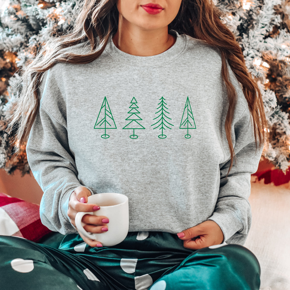 A woman wearing a sport gray Gildan crewneck sweatshirt with 4 line drawn Christmas trees on it.