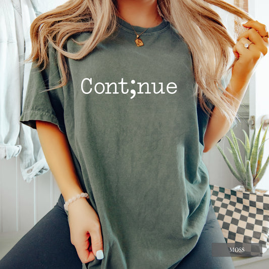 A woman wearing an oversized sage green t-shirt with the word 'Continue' printed in white across the chest, styled with black jeans in a bright indoor setting.