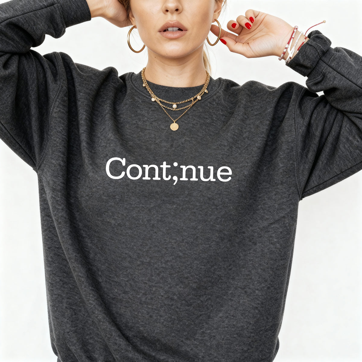 A woman wearing a dark heather gray crewneck sweatshirt with the word 'Continue' printed in white serif text across the chest. She is wearing layered gold necklaces, hoop earrings, and bracelets against a clean white background.