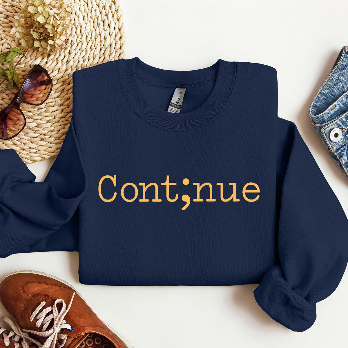 A navy blue sweatshirt with the word 'Continue' printed in yellow across the chest, styled in a flat lay with a straw hat, sunglasses, brown shoes, and denim on a white surface.