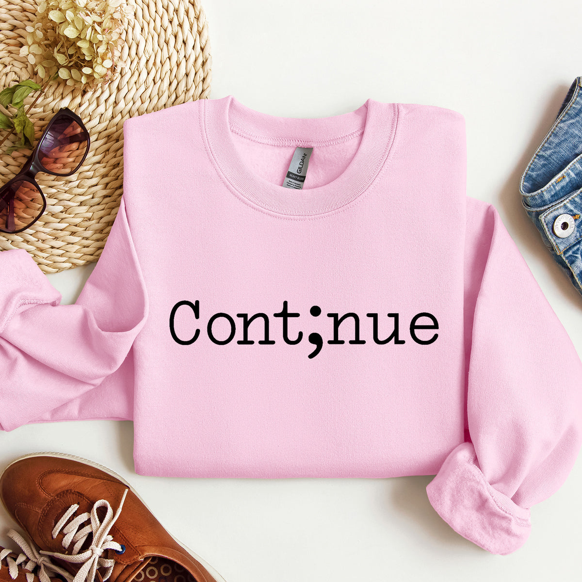 A folded light pink crewneck sweatshirt with the word 'Continue' printed in black across the chest, styled in a flat lay with sunglasses, a woven hat, a brown shoe, and a denim item on a white background.