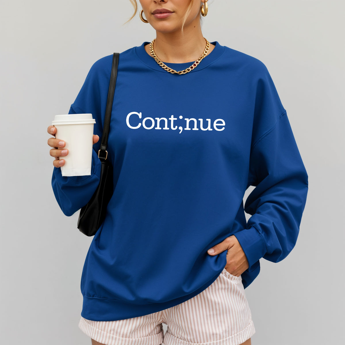 A woman wearing a royal blue crewneck sweatshirt with the word 'Continue' printed in white across the chest, holding a white coffee cup and wearing pink-and-white striped shorts against a light gray background.