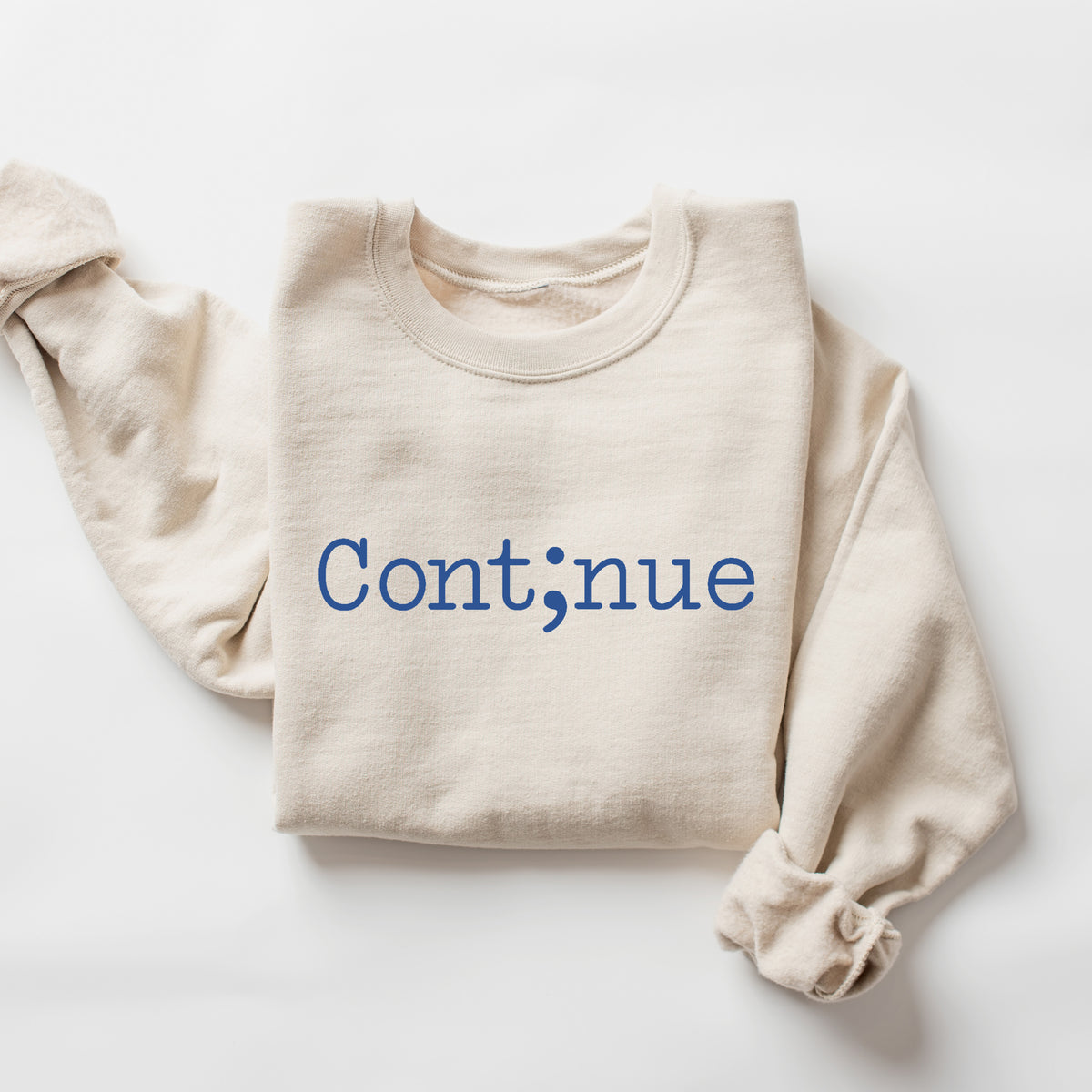 A folded cream-colored long-sleeve sweatshirt with the word 'Continue' printed in blue across the chest, featuring a semicolon as the letter 'n'. The sweatshirt is laid flat on a clean white background.