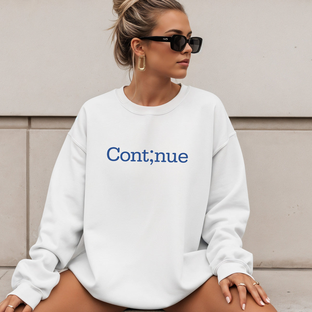 A woman wearing an oversized white crewneck sweatshirt with the word 'Cont;inue' printed in blue on the chest, posed against a neutral concrete wall.