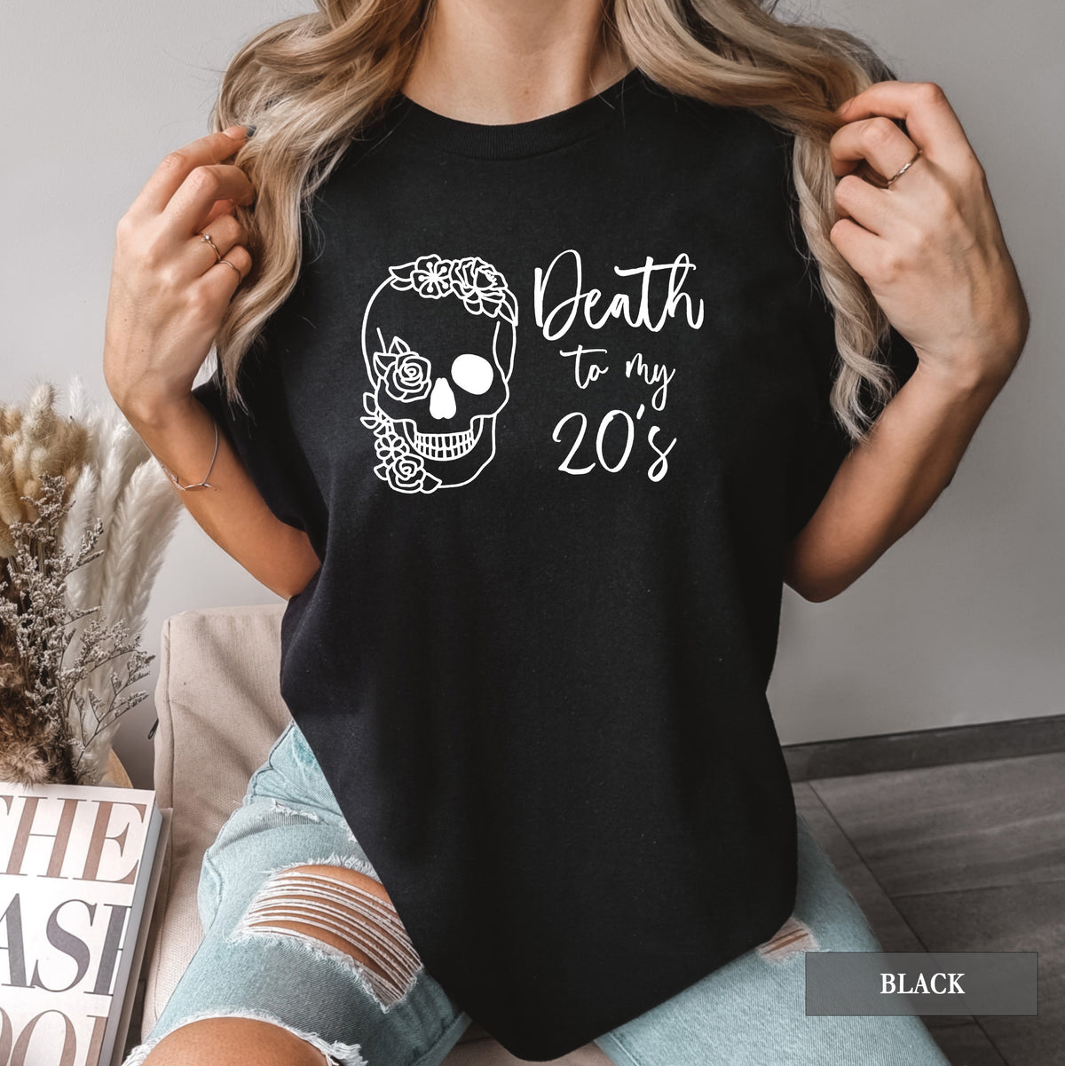 A woman wearing a black graphic t-shirt with a white sugar skull and the text 'Death to my 20's'. She is holding her hair and wearing distressed denim shorts in a cozy indoor setting.