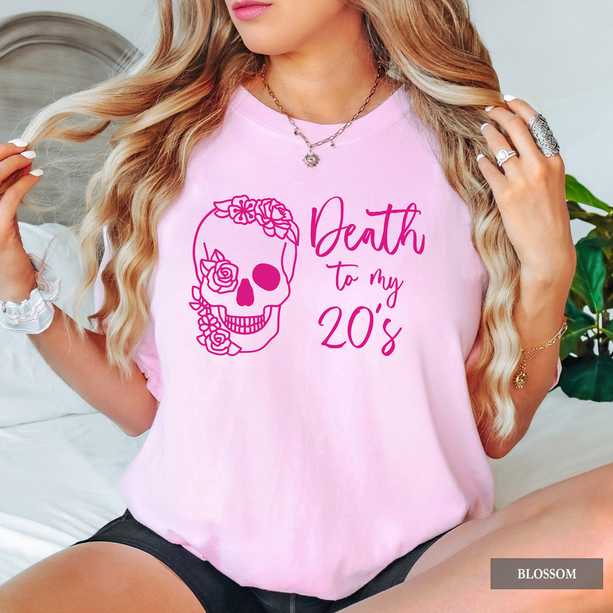 A woman wearing a light pink graphic t-shirt with a dark pink skull and roses design and the text 'Death to my 20's'.