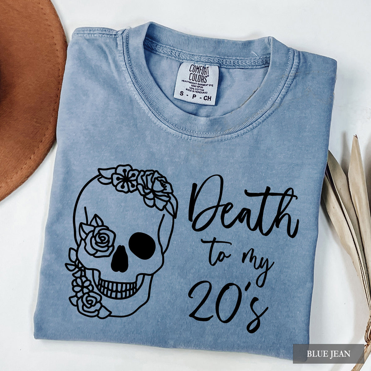 A folded light blue t-shirt with a black graphic of a floral skull and the text 'Death to my 20's'. The shirt is displayed on a white surface with a brown hat and dried leaves as props.