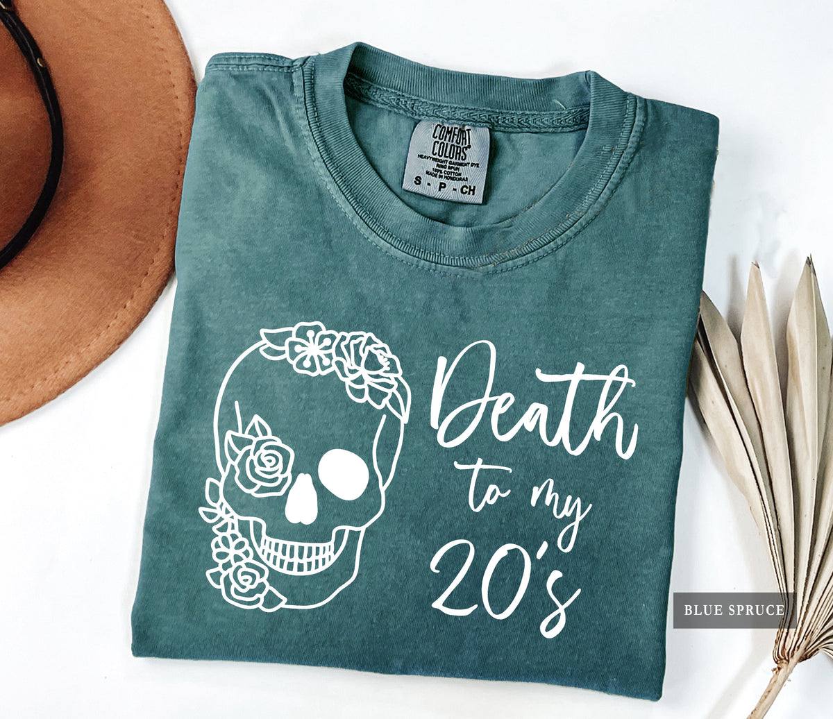 A folded slate blue t-shirt with a white graphic of a skull decorated with flowers and the text 'Death to my 20's'. The shirt is styled in a flat-lay composition on a white background, partially covered by a brown hat on the left and dried palm leaves on the right.