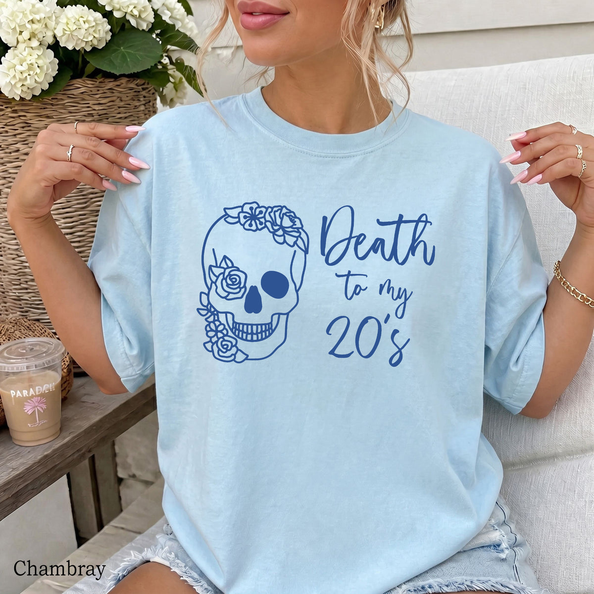 A woman wearing a light chambray blue graphic t-shirt with a dark blue floral skull and the text 'Death to my 20's'. She is sitting on a white outdoor sofa next to a table with a drink.