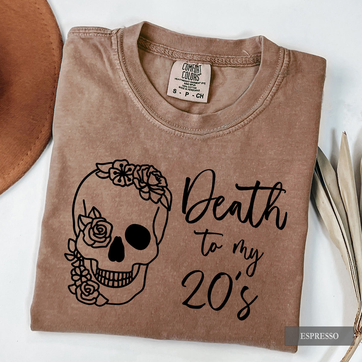 A folded brown t-shirt with a black graphic of a skull decorated with flowers and the text 'Death to my 20's'. The shirt is displayed on a white background with a brown hat edge and dried leaves as props.