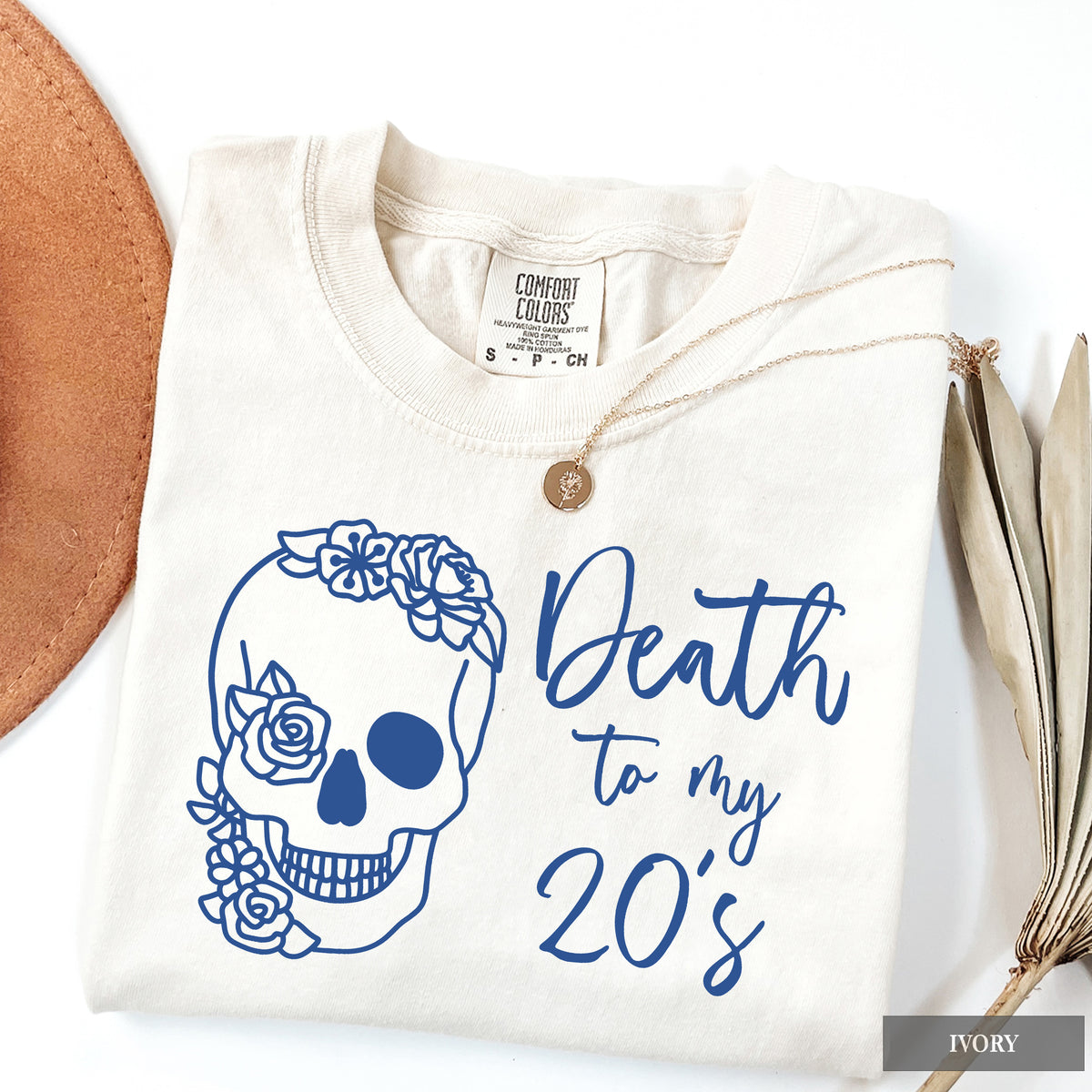 A folded cream-colored t-shirt with a blue graphic of a skull decorated with flowers and the text 'Death to my 20's'. The shirt is styled with a wooden pendant necklace, dried leaves, and a brown leather circle on a white background.