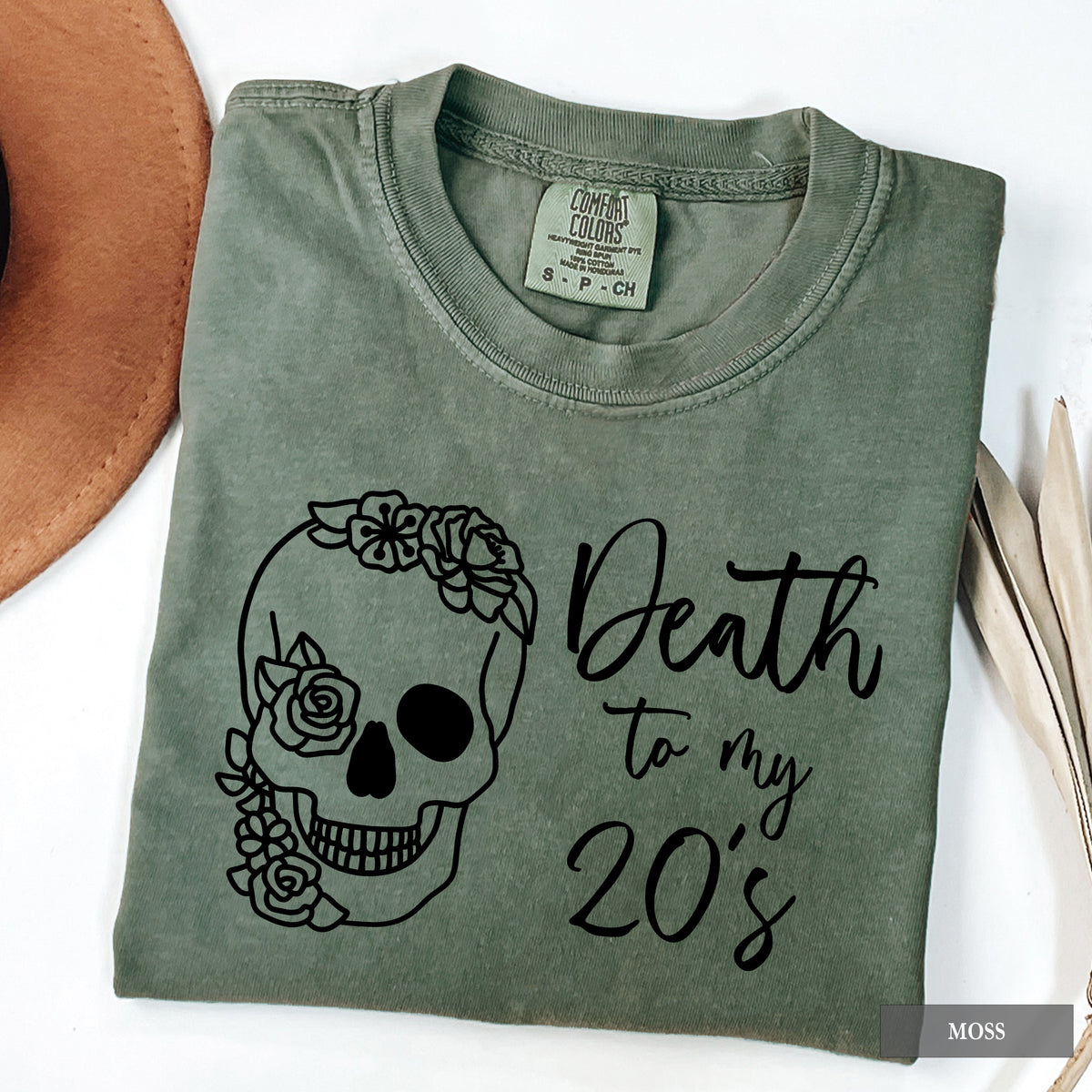 An olive green folded t-shirt with a black graphic of a floral sugar skull and the text 'Death to my 20's'. The shirt is styled on a white background with a brown hat on the left and dried leaves on the right.