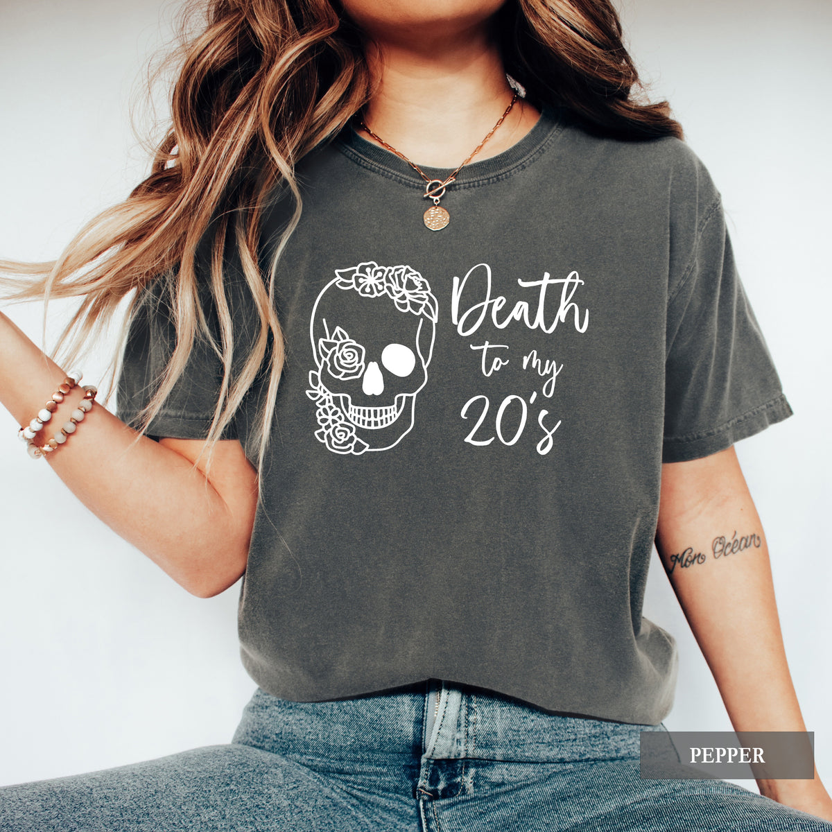 A woman wearing a faded dark gray graphic t-shirt with a white sugar skull and the text 'Death to my 20's'. The t-shirt has a warm, washed texture.