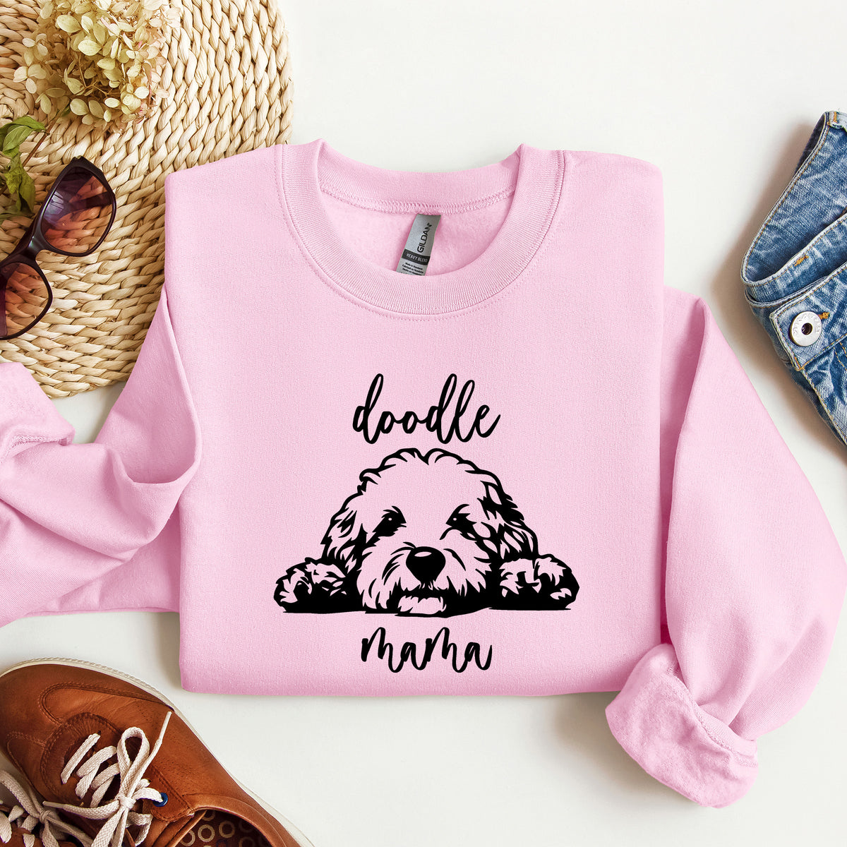 A folded light pink crewneck sweatshirt with a black graphic of a doodle dog and the words 'doodle mama' on the front, styled in a flat lay with sunglasses, a woven hat, jeans, and a brown shoe.