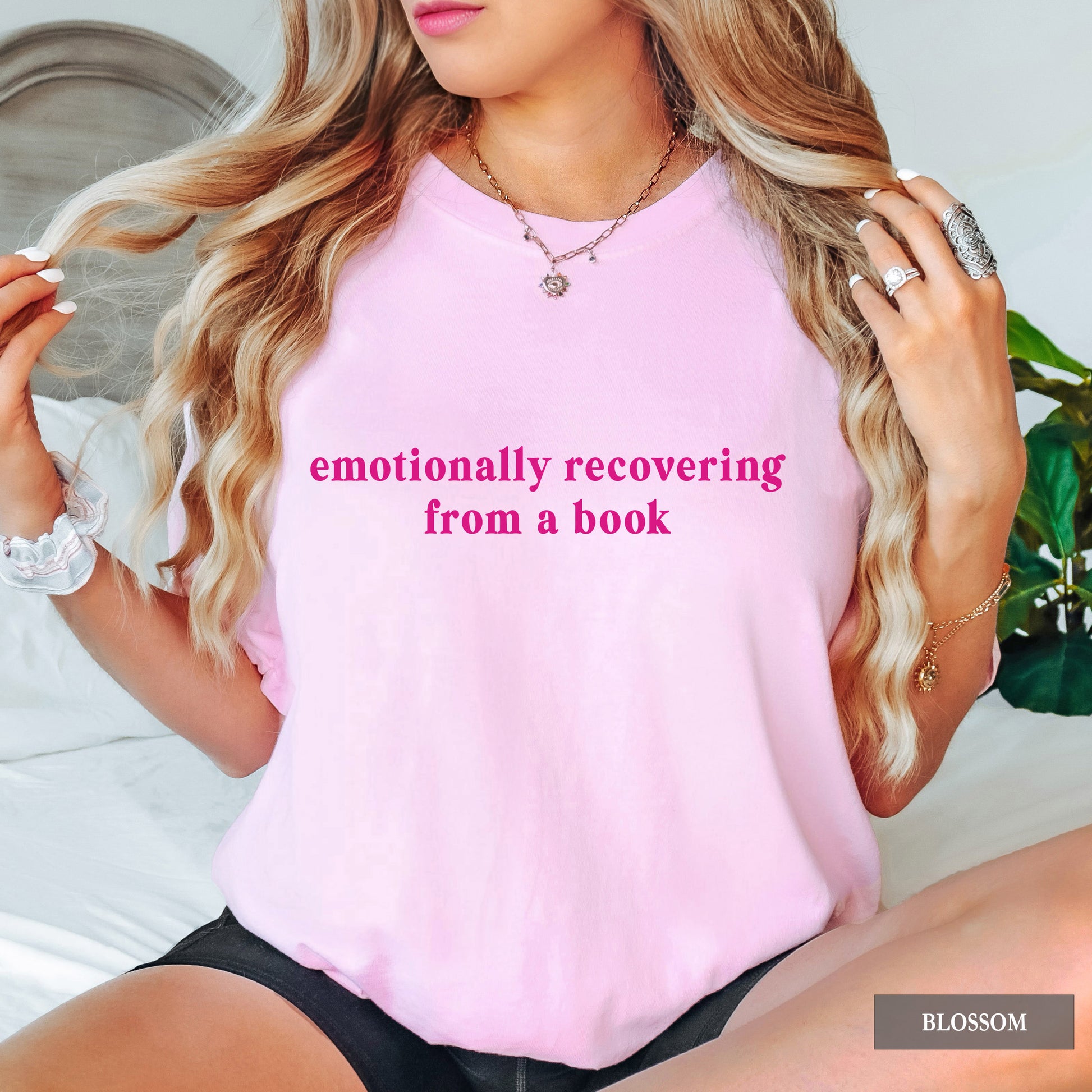 A woman wearing a light pink t-shirt with 'emotionally recovering from a book' printed in pink text on the front. She is sitting on a bed with a headboard and a green plant visible in the background.