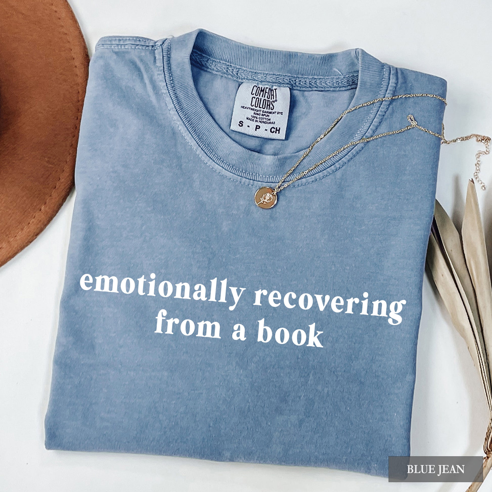A folded light blue t-shirt with white text reading 'emotionally recovering from a book' on the front, styled on a white background with a brown hat edge, a necklace, and dried leaves. A 'COMFORT COLORS' neck label is visible, and a 'BLUE JEAN' color label appears in the bottom right corner.