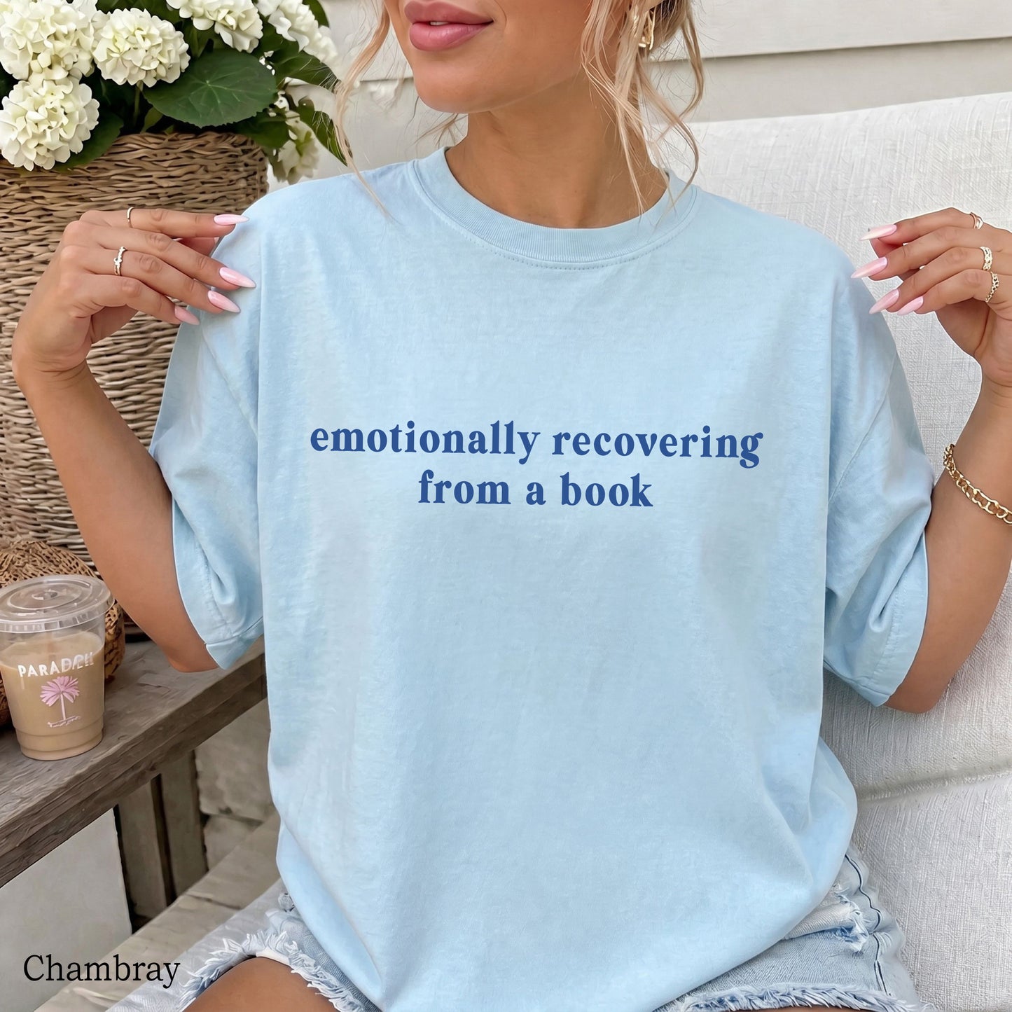 A woman wearing a light chambray blue oversized T-shirt with the text 'emotionally recovering from a book' printed in dark blue across the chest. She is sitting on a white couch with a coffee cup and flowers in the background.