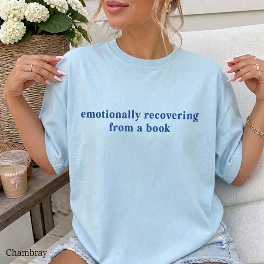 A woman wearing a light chambray blue oversized T-shirt with the text 'emotionally recovering from a book' printed in dark blue across the chest. She is sitting on a white couch with a coffee cup and flowers in the background.