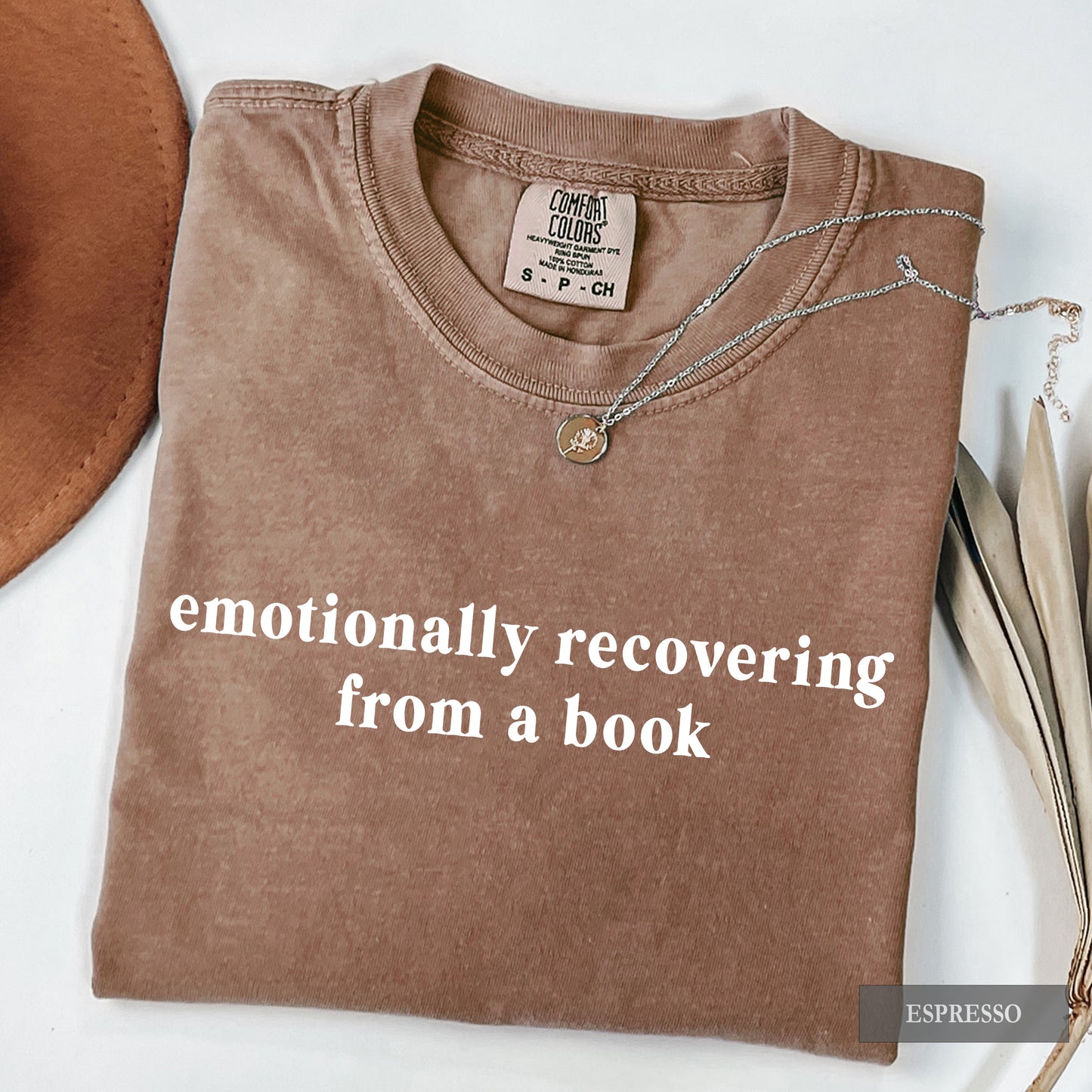 A folded espresso brown t-shirt with the text 'emotionally recovering from a book' printed in white. The shirt is styled with a silver necklace and a brown hat on a white background.