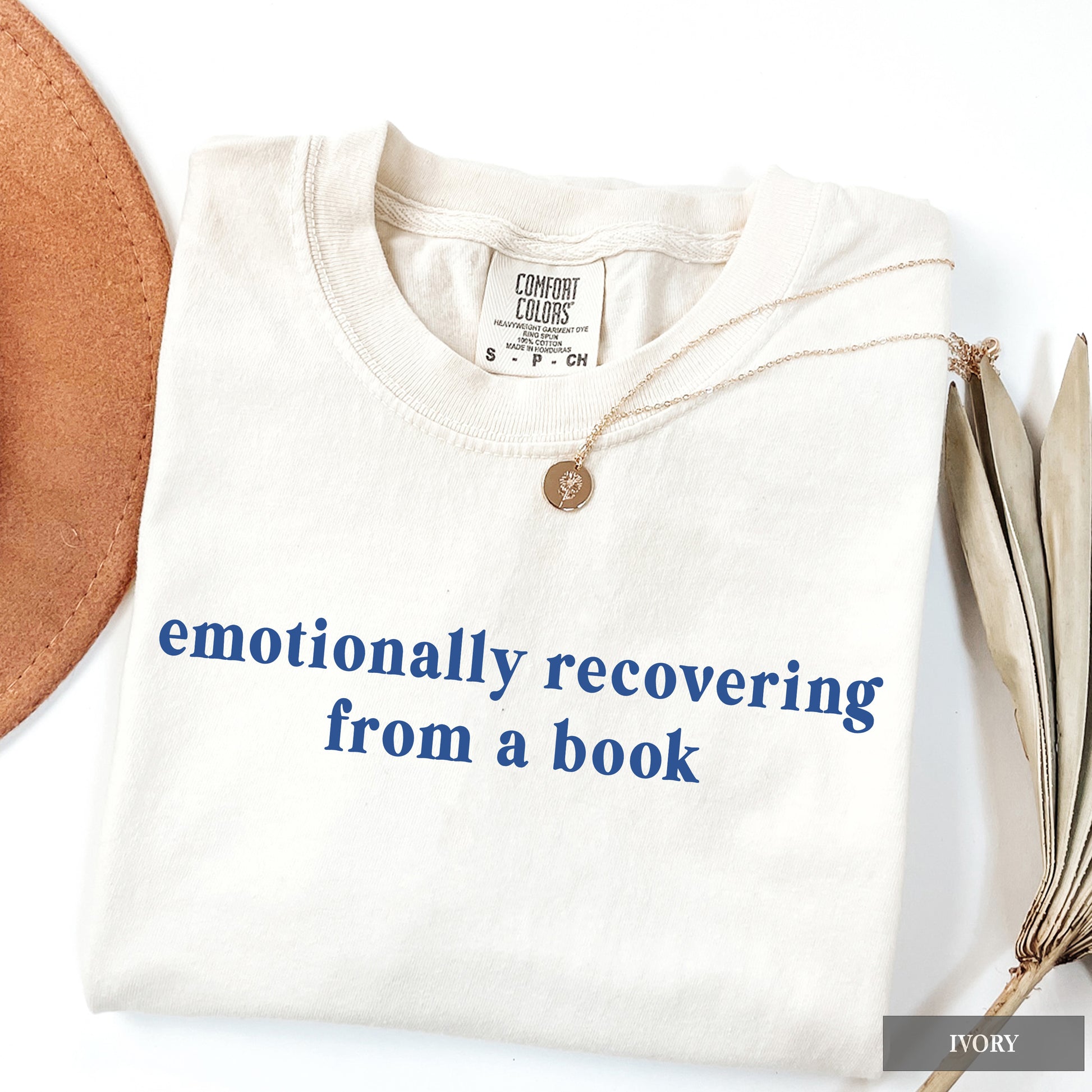 A folded ivory crewneck t-shirt with the phrase 'emotionally recovering from a book' printed in blue across the chest. The shirt is styled with a gold pendant necklace and surrounded by decorative props, including a leather circle on the left and dried leaves on the right, on a white background.