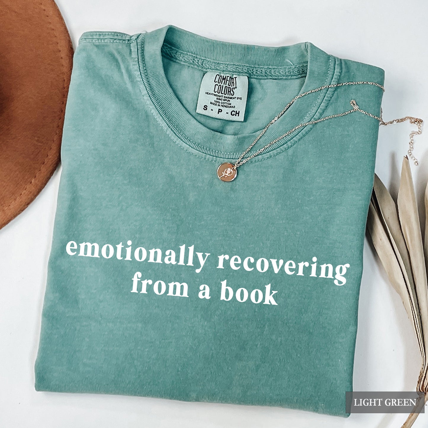 A folded light green t-shirt with the phrase 'emotionally recovering from a book' printed in white lowercase letters across the front. The shirt is styled with a delicate silver necklace featuring a round bronze pendant, a brown leather circle on the left, and dried leaves on the right, all on a white background.