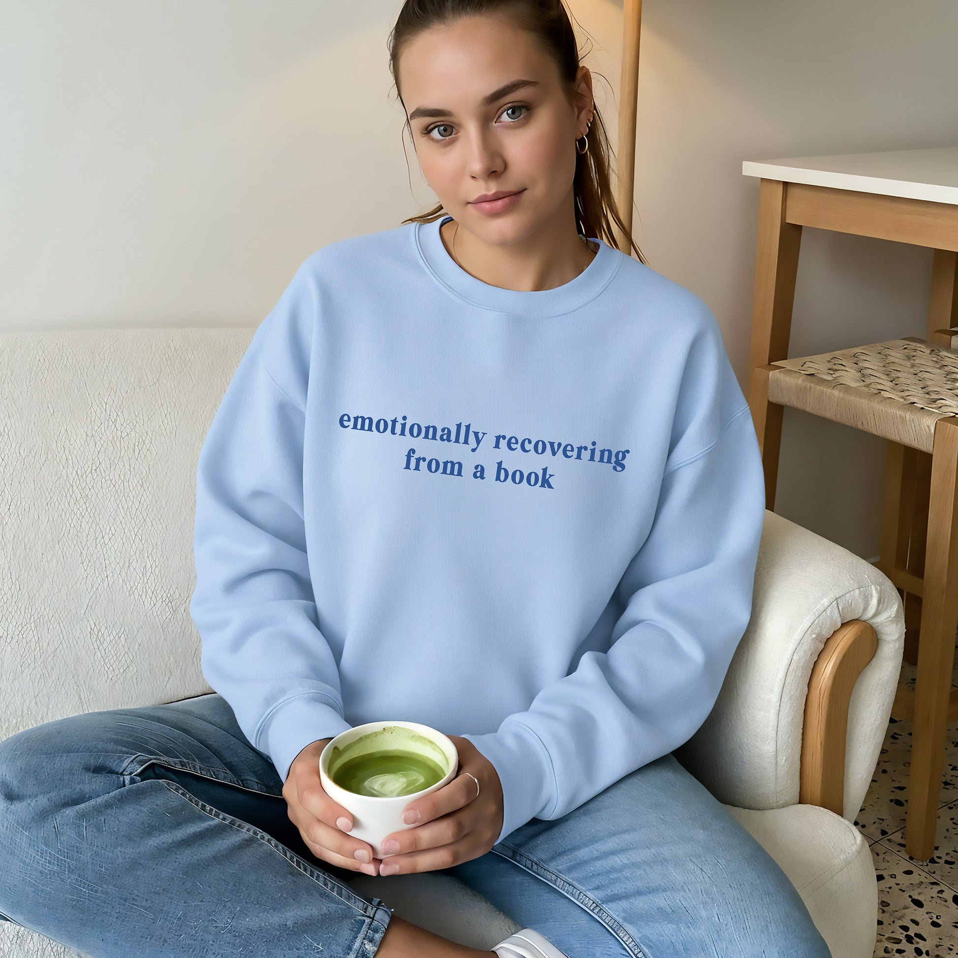 A woman sits on a white armchair holding a cup of green matcha tea, wearing a light blue crewneck sweatshirt with the text 'emotionally recovering from a book' printed in blue on the chest.