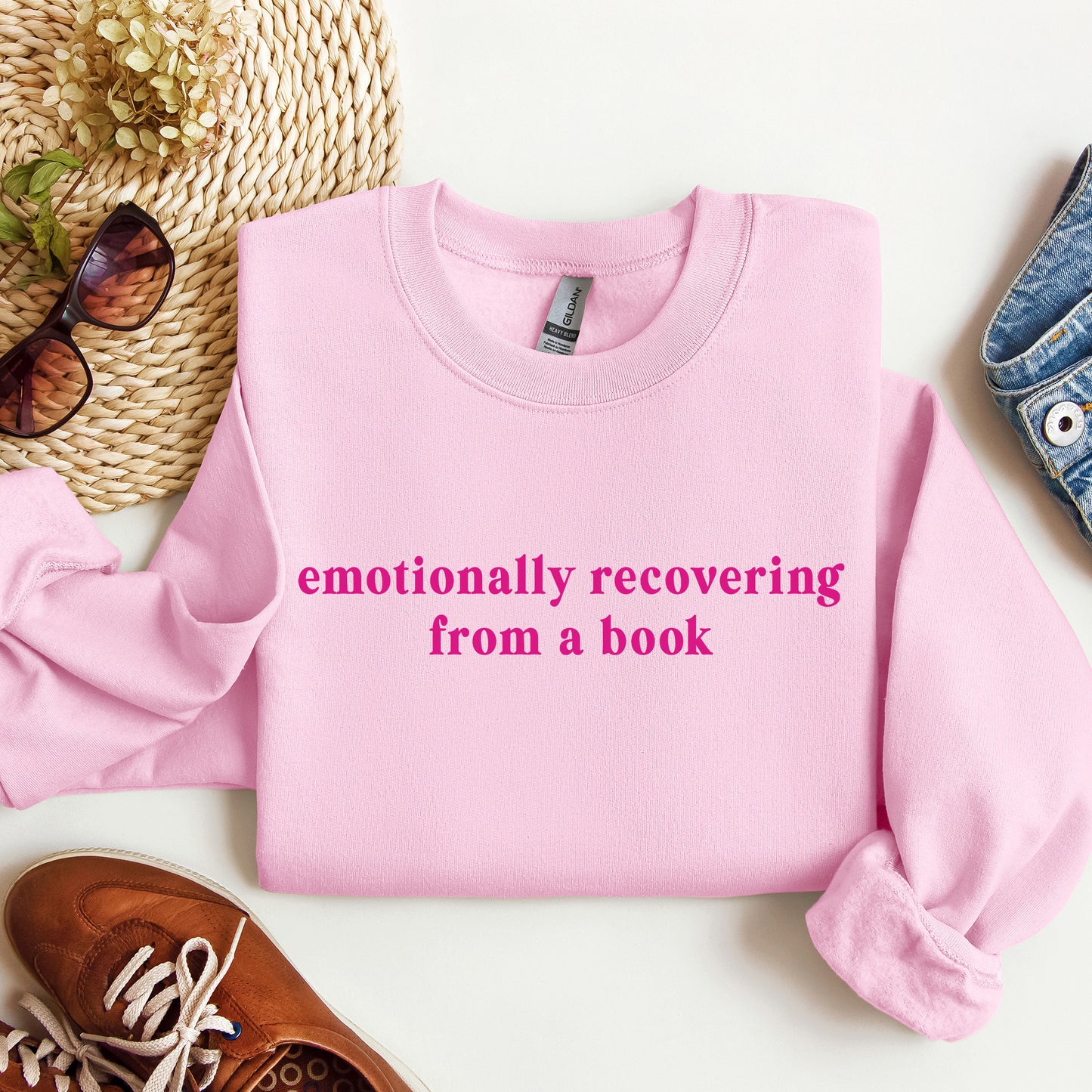 A folded light pink crewneck sweatshirt with the phrase 'emotionally recovering from a book' printed in bright pink across the chest. The sweatshirt is styled in a flat lay with a woven hat, sunglasses, a brown shoe, and a denim item on a white background.