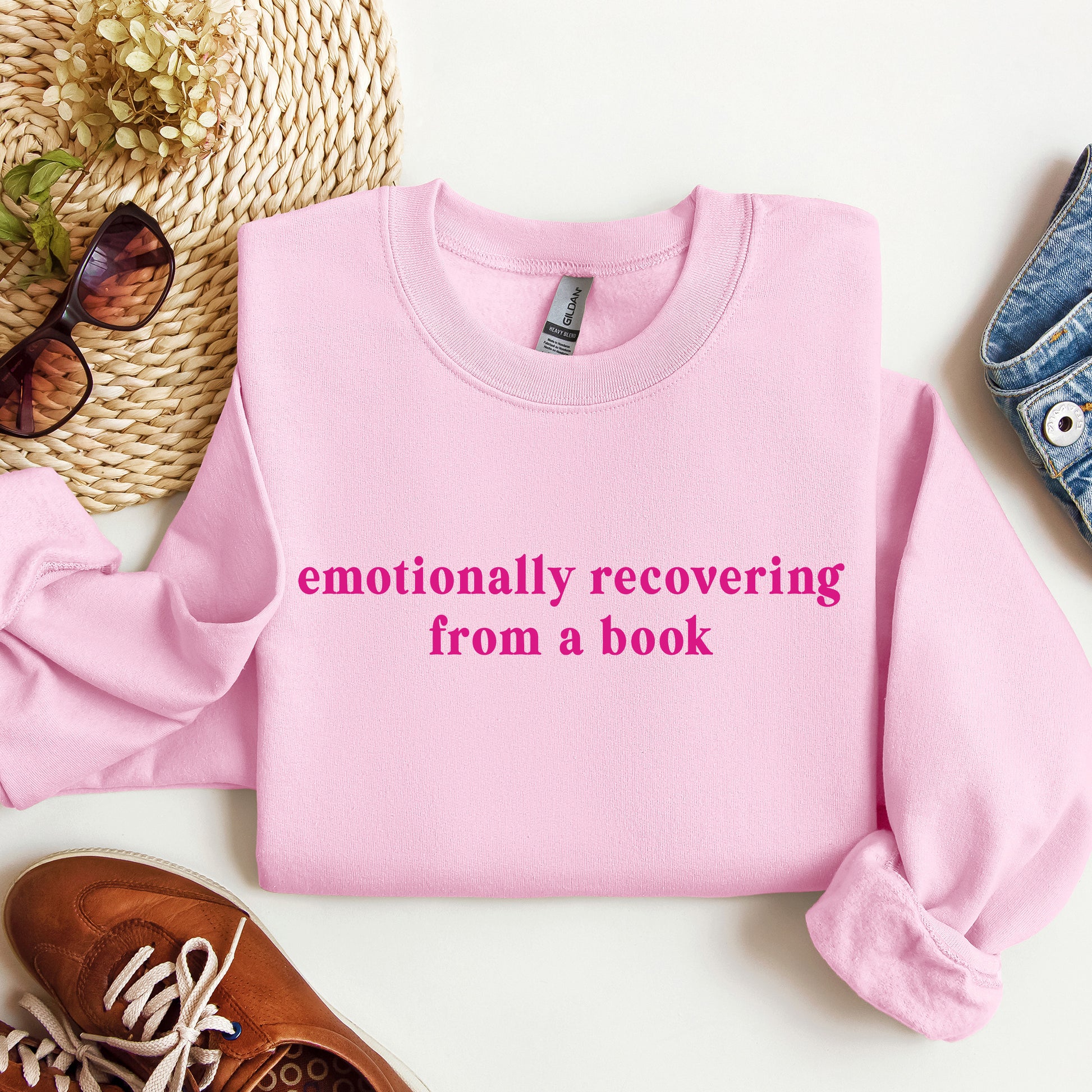 A folded light pink crewneck sweatshirt with the phrase 'emotionally recovering from a book' printed in bright pink across the chest. The sweatshirt is styled in a flat lay with a woven hat, sunglasses, a brown shoe, and a denim item on a white background.
