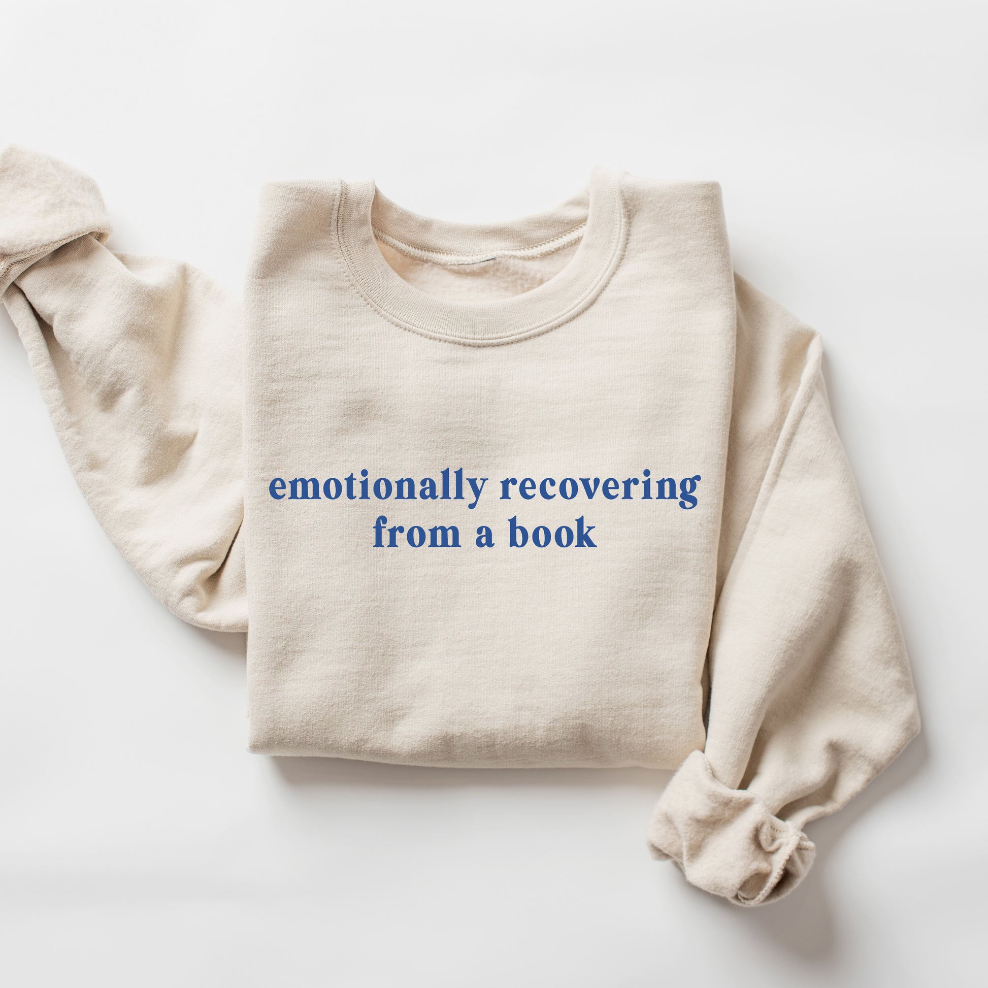 A folded cream-colored crewneck sweatshirt with the phrase 'emotionally recovering from a book' printed in blue lowercase serif text across the chest, displayed on a clean white background.
