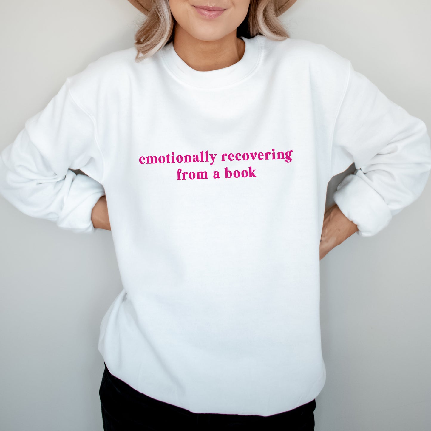 A person wearing a white crewneck sweatshirt with the phrase 'emotionally recovering from a book' printed in pink lowercase letters across the chest, posed against a light gray background.