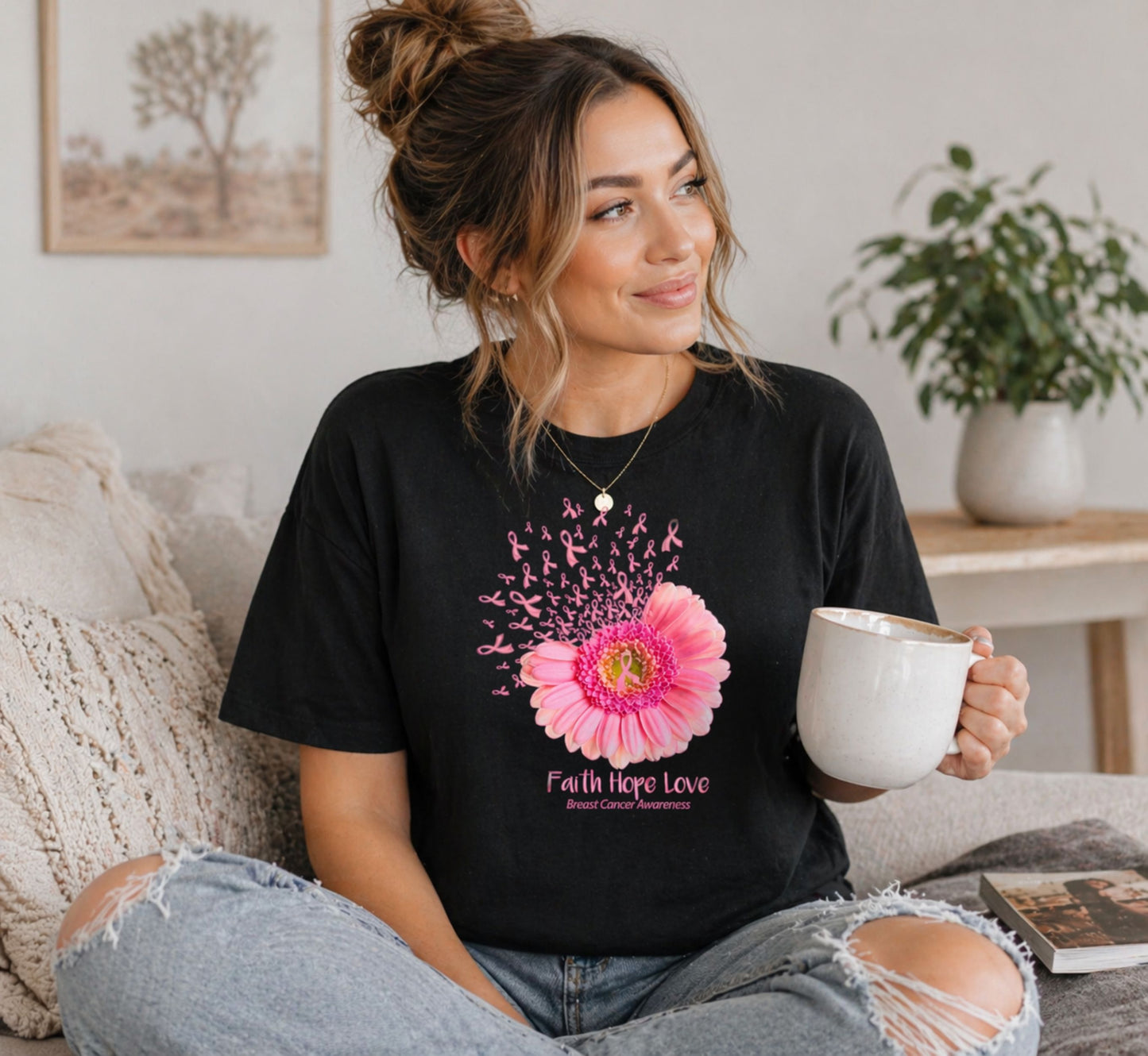Woman wearing a black t-shirt with a pink flower breast cancer awareness design and text, holding a white mug in a cozy living room.