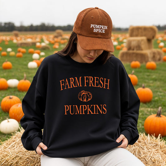 A woman sitting on a hay bale in a pumpkin field, wearing a black sweatshirt with orange text reading 'FARM FRESH PUMPKINS' and a brown corduroy cap with 'PUMPKIN SPICE' embroidered on it.