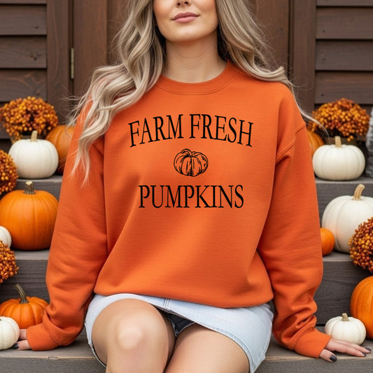 A woman wearing an orange crewneck sweatshirt with 'FARM FRESH PUMPKINS' printed in black text and a pumpkin graphic, posed on wooden steps surrounded by pumpkins.