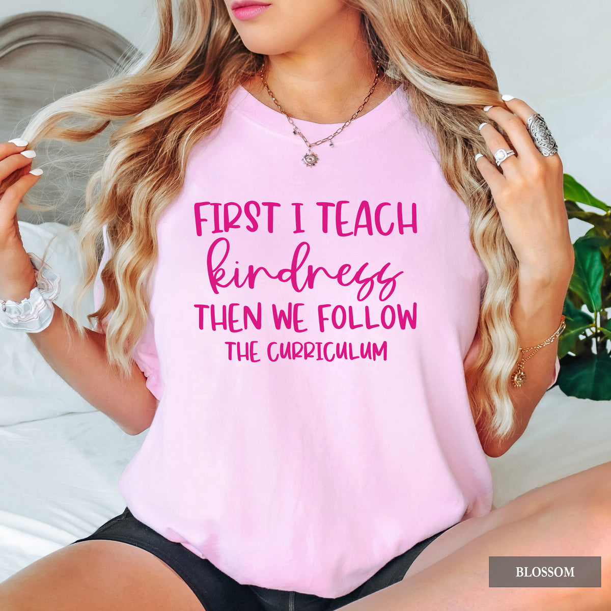 A woman wearing a light pink t-shirt with 'First I Teach Kindness' printed in pink text on the front. She is sitting on a bed with a headboard and a green plant visible in the background.