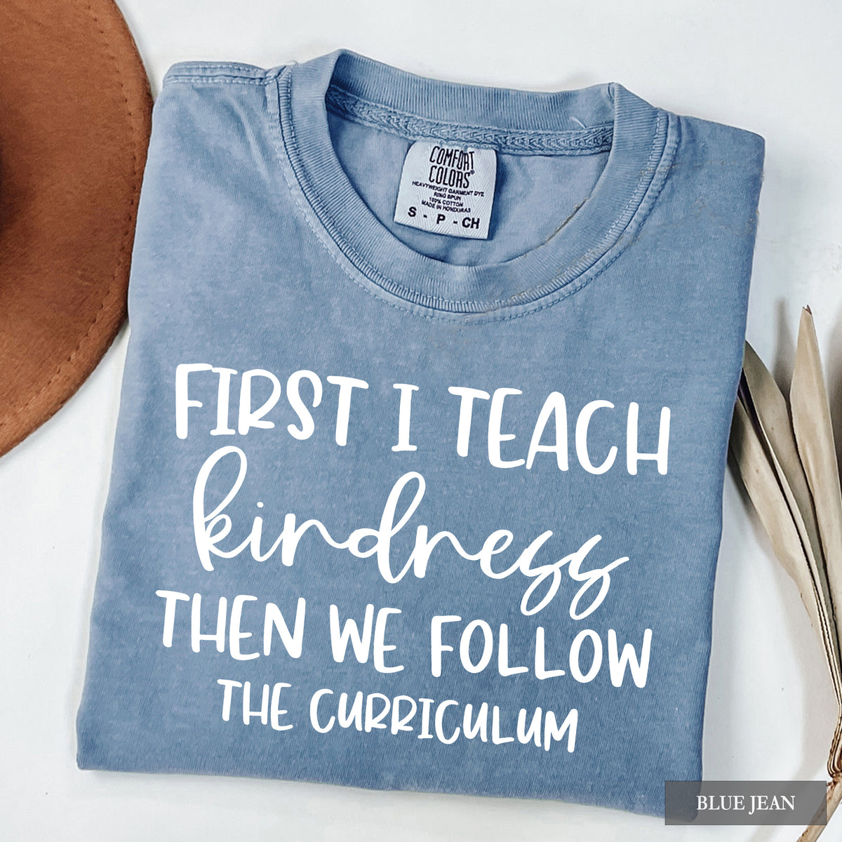 A folded light blue t-shirt with white text reading 'FIRST I TEACH kindness THEN WE FOLLOW THE CURRICULUM' on a white background, styled with a brown hat and dried leaves. The neck label shows 'Comfort Colors' and 'S-PCH'.