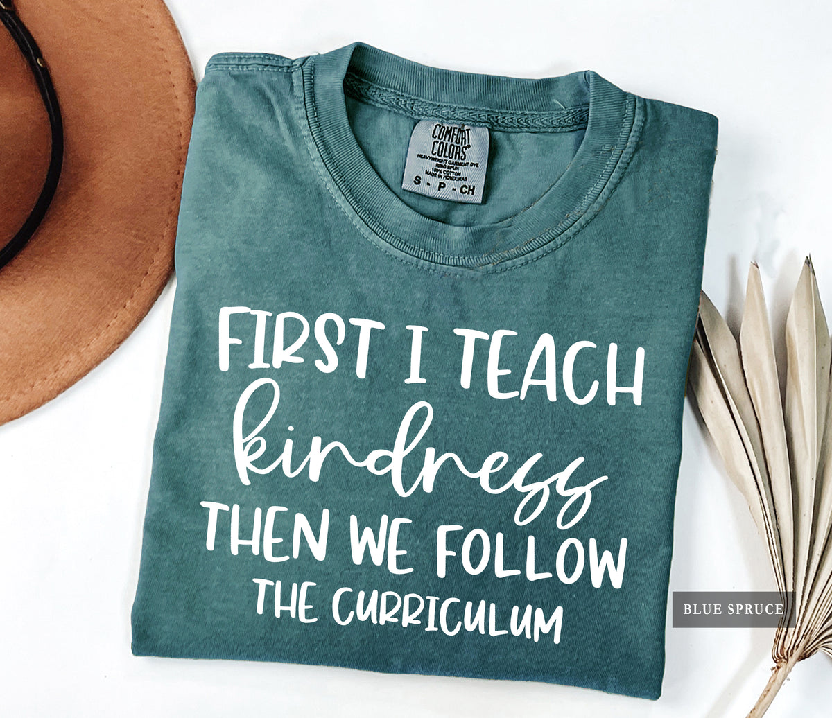 A folded slate blue-green t-shirt with white text reading 'FIRST I TEACH kindness THEN WE FOLLOW THE CURRICULUM' is styled in a flat lay with a brown hat and dried palm leaves on a white background. The tag reads 'Comfort Colors' and a watermark in the corner reads 'BLUE SPRUCE'.