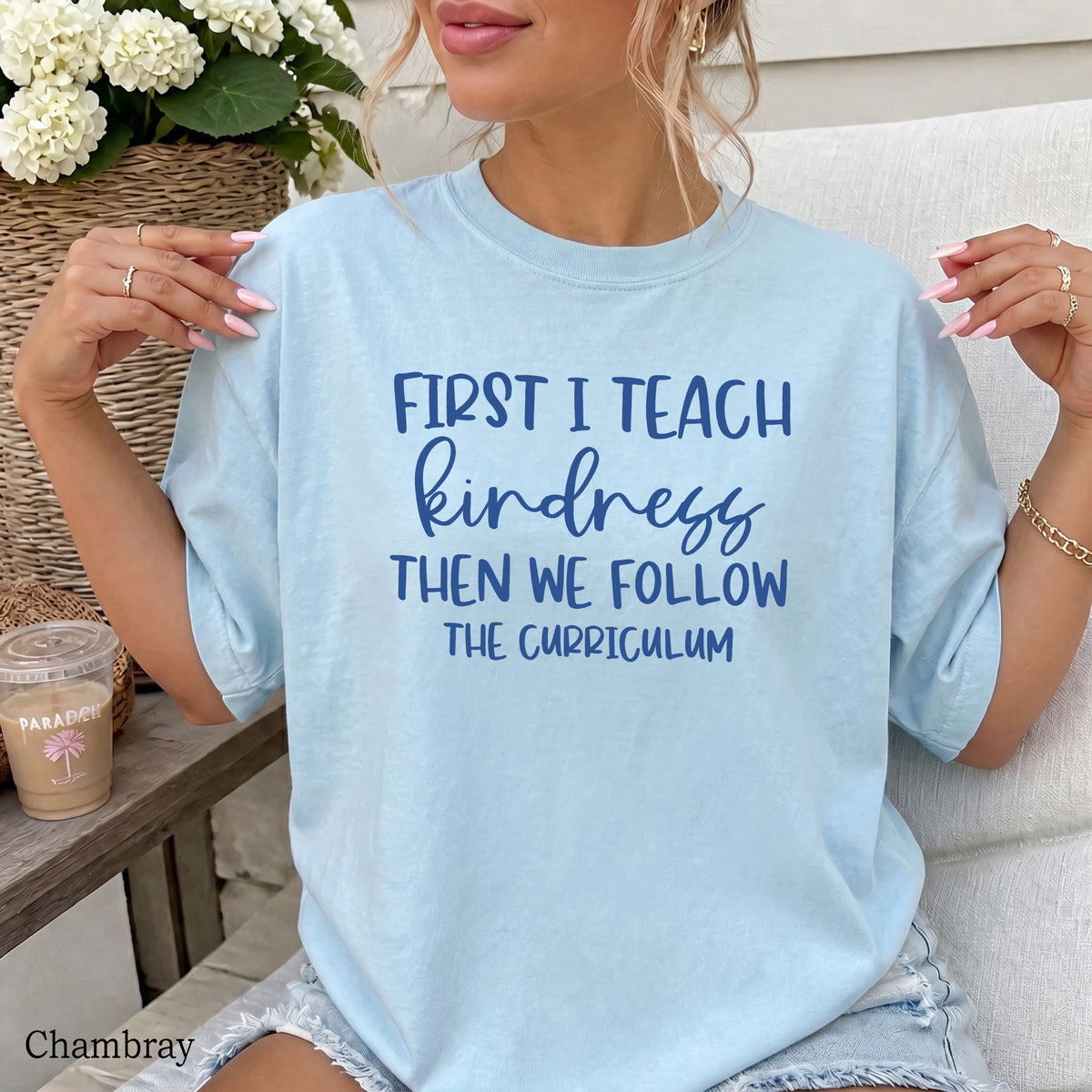 A woman wearing a light chambray blue t-shirt with the text 'FIRST I TEACH kindness THEN WE FOLLOW THE CURRICULUM' printed in dark blue on the front. She is sitting on a white couch with a coffee cup and flowers in the background.