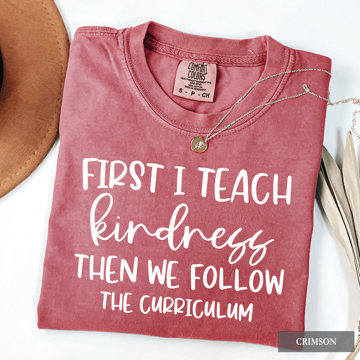 A folded dusty rose/crimson t-shirt with white text reading 'FIRST I TEACH kindness THEN WE FOLLOW THE CURRICULUM' on a white background, styled with a necklace, a brown hat edge, and dried leaves. A 'CRIMSON' color label is visible in the corner.