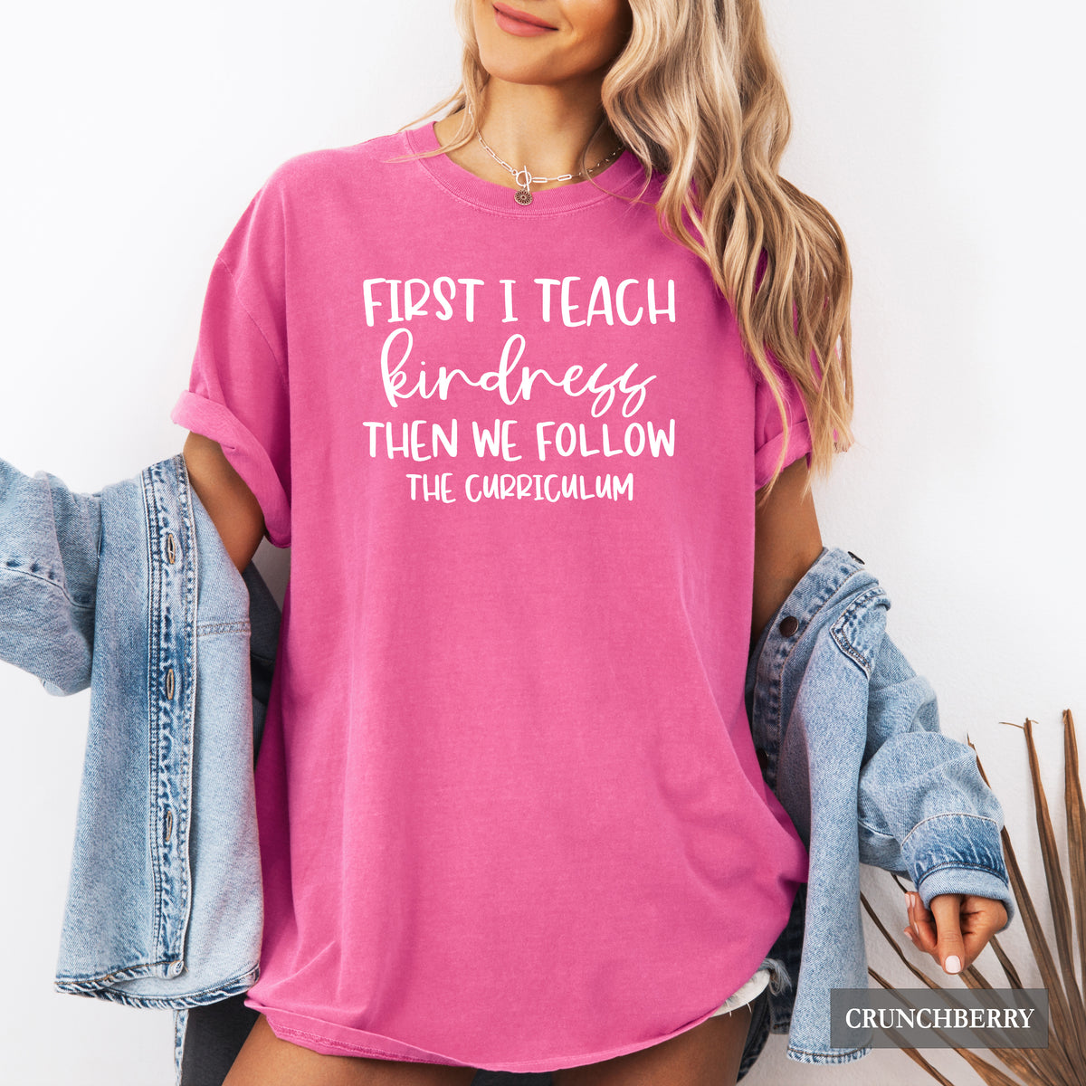 A woman wearing a bright pink graphic t-shirt with white text that reads 'FIRST I TEACH kindness THEN WE FOLLOW THE CURRICULUM'. She has a light-wash denim jacket draped off her shoulders against a clean white wall.