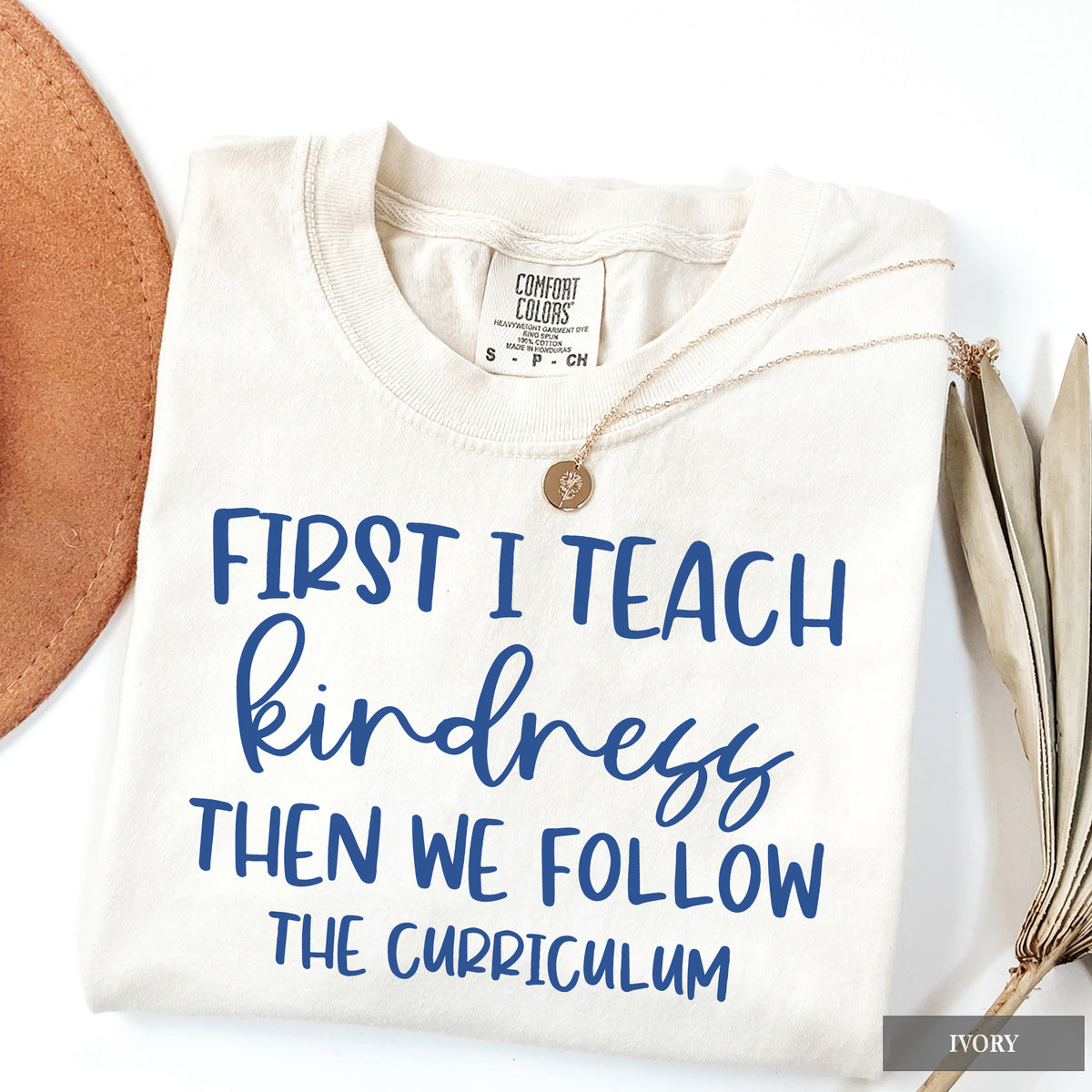 An ivory folded t-shirt with blue text reading 'FIRST I TEACH kindness THEN WE FOLLOW THE CURRICULUM', styled with a gold coin necklace and dried palm leaves on a white background.