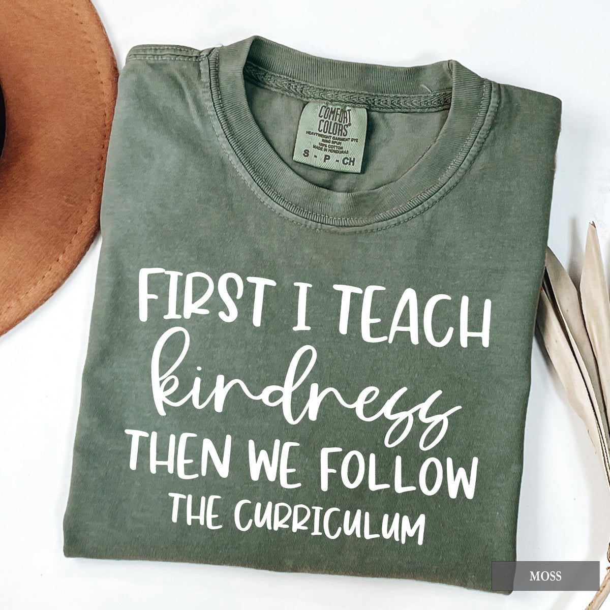 An olive green folded t-shirt with white printed text reading 'FIRST I TEACH kindness THEN WE FOLLOW THE CURRICULUM'. The t-shirt is styled in a flat-lay arrangement with a brown hat on the left and dried leaves on the right, on a white background.