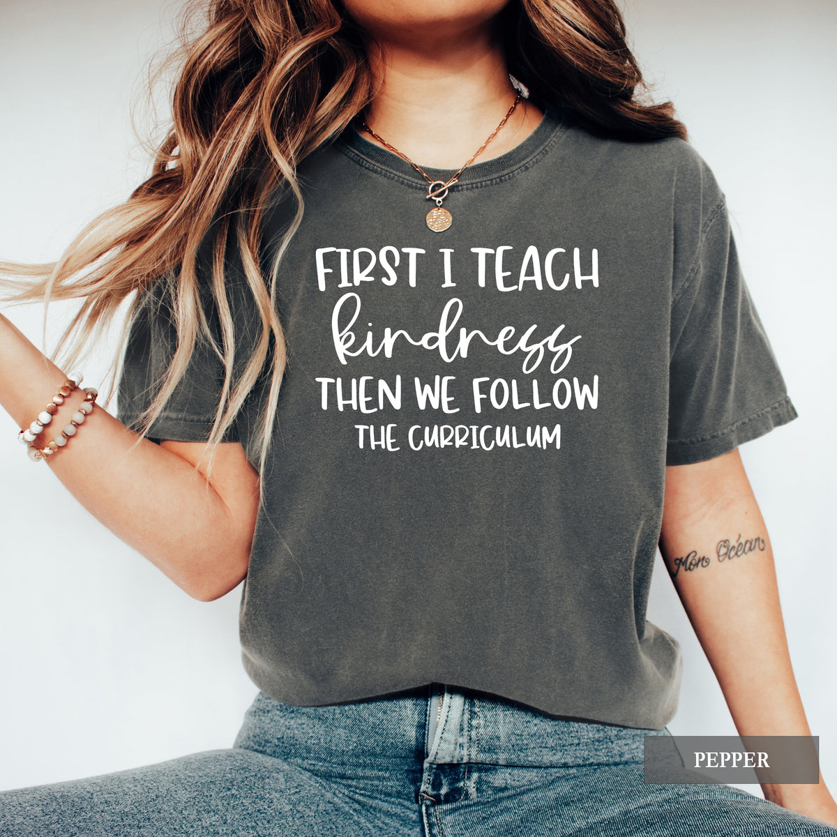 A woman wearing a dark gray washed graphic t-shirt with the text 'FIRST I TEACH kindness THEN WE FOLLOW THE CURRICULUM' in white. She is also wearing blue jeans, a gold necklace, and bracelets, set against a plain light gray background.
