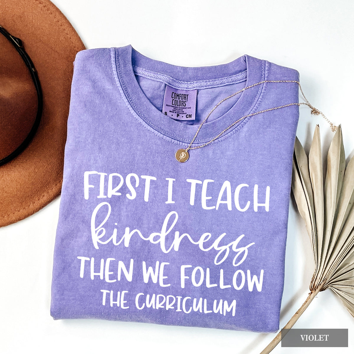 A folded lavender-purple t-shirt with white text reading 'FIRST I TEACH kindness THEN WE FOLLOW THE CURRICULUM' is styled on a white background with a brown hat, a gold necklace, and dried palm leaves. A 'VIOLET' brand label is visible on the collar.