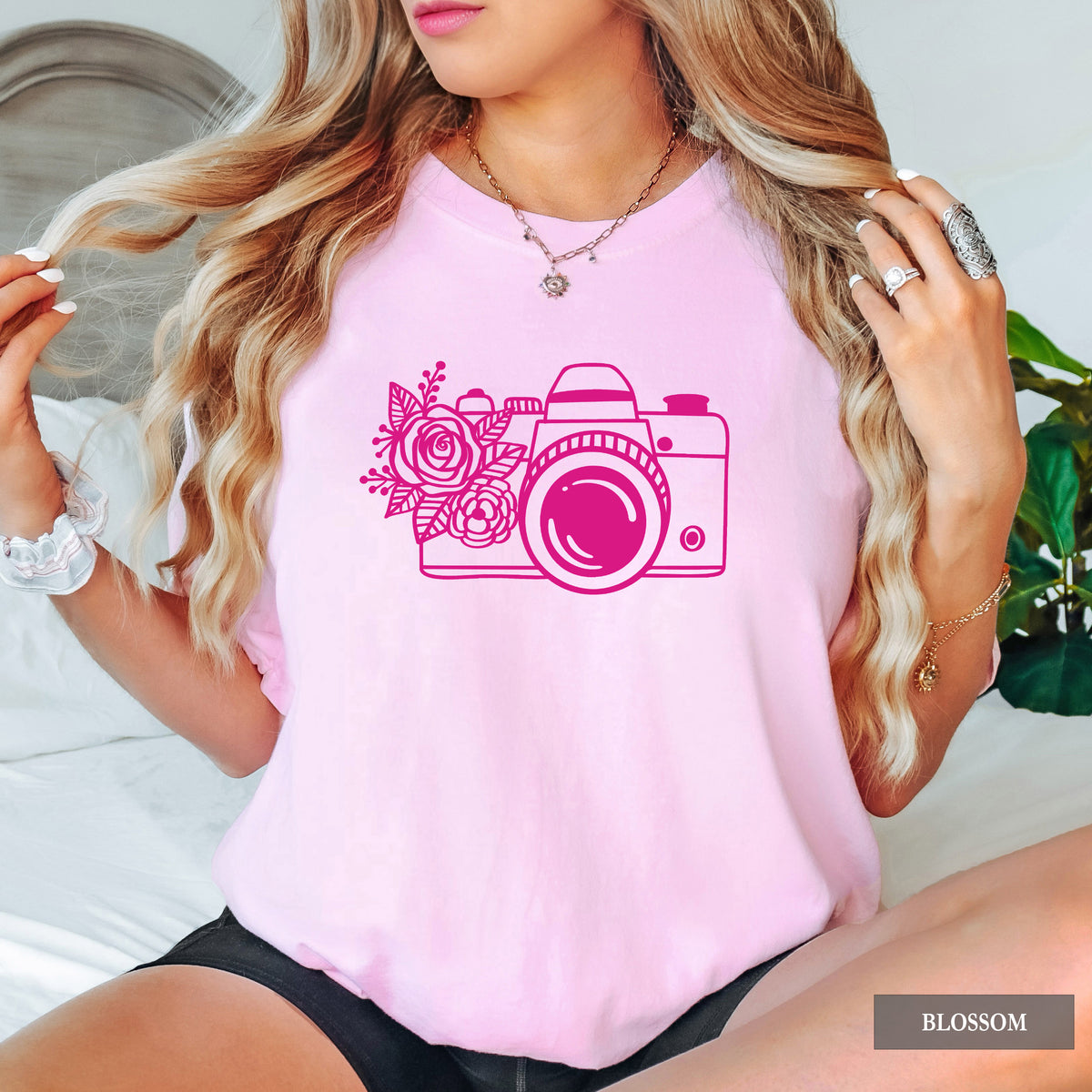 A woman wearing a light pink short-sleeve t-shirt with a dark pink graphic of a camera decorated with flowers. The image is a lifestyle shot with a 'BLOSSOM' color label in the bottom right corner.