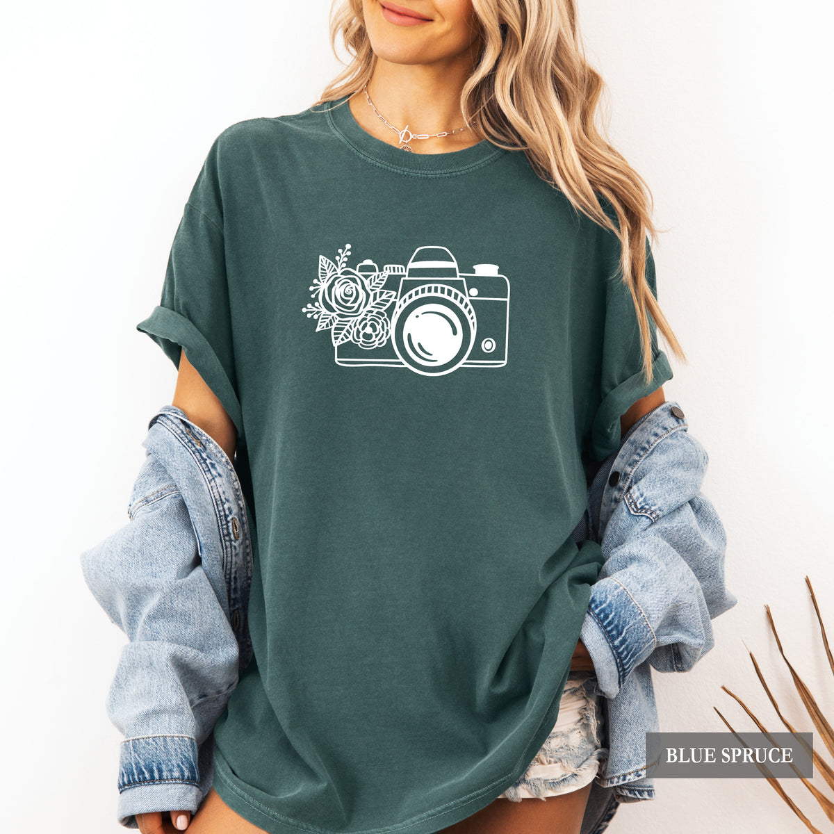 A woman wearing an oversized dark teal-green t-shirt with a white camera and floral graphic on the front, styled with rolled-up light-wash denim sleeves and denim shorts. A 'BLUE SPRUCE' brand label is visible in the bottom right corner.
