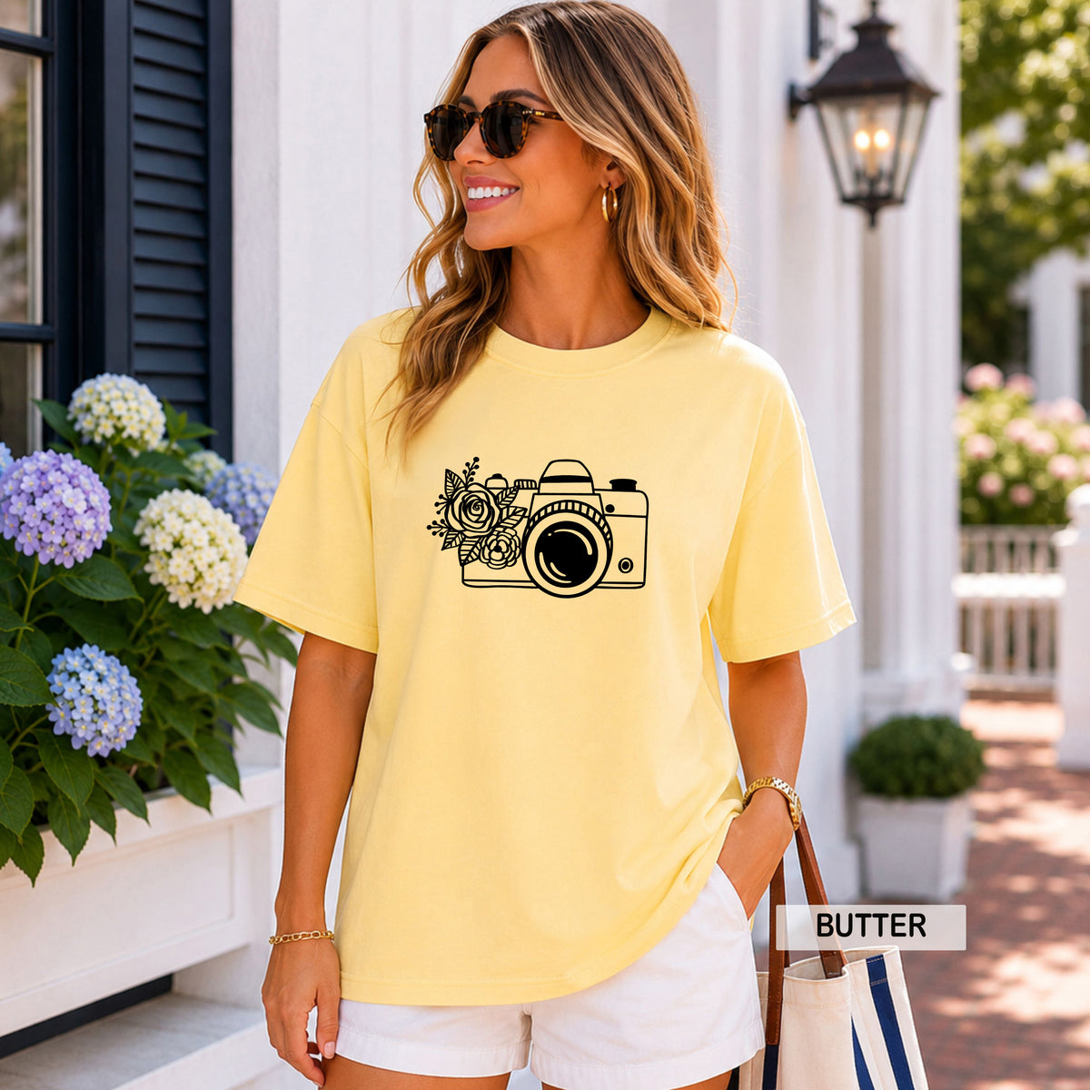 A woman wearing a pale yellow graphic t-shirt with a black camera and floral print, styled with white shorts in an outdoor porch setting.