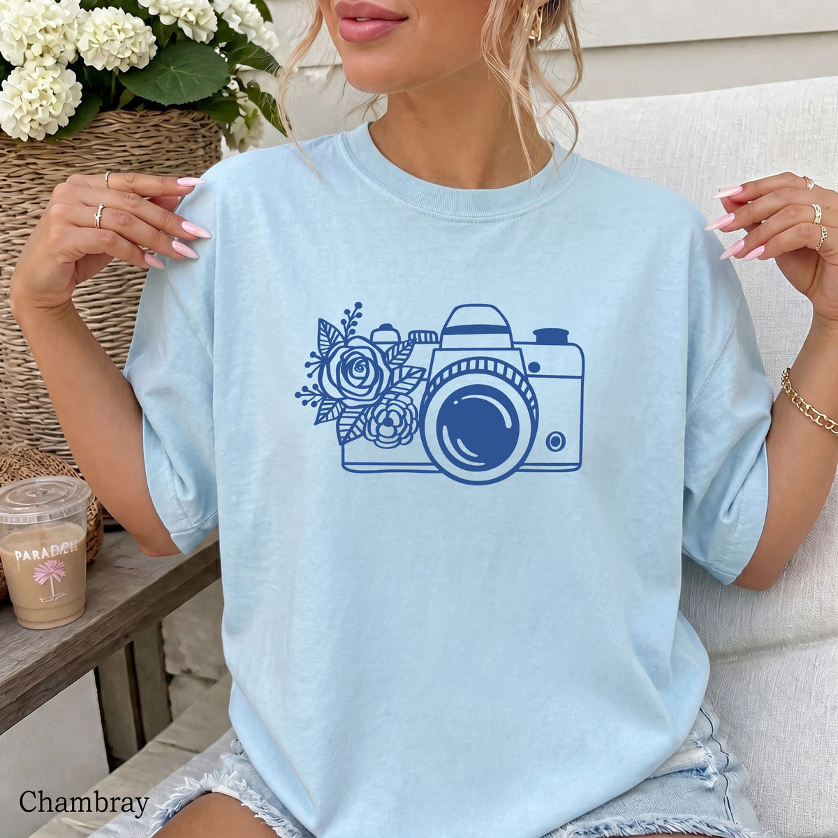 A woman wearing a light chambray blue oversized T-shirt with a dark blue graphic of a camera and flowers on the front, seated on a white chair with a coffee cup and flowers in the background.