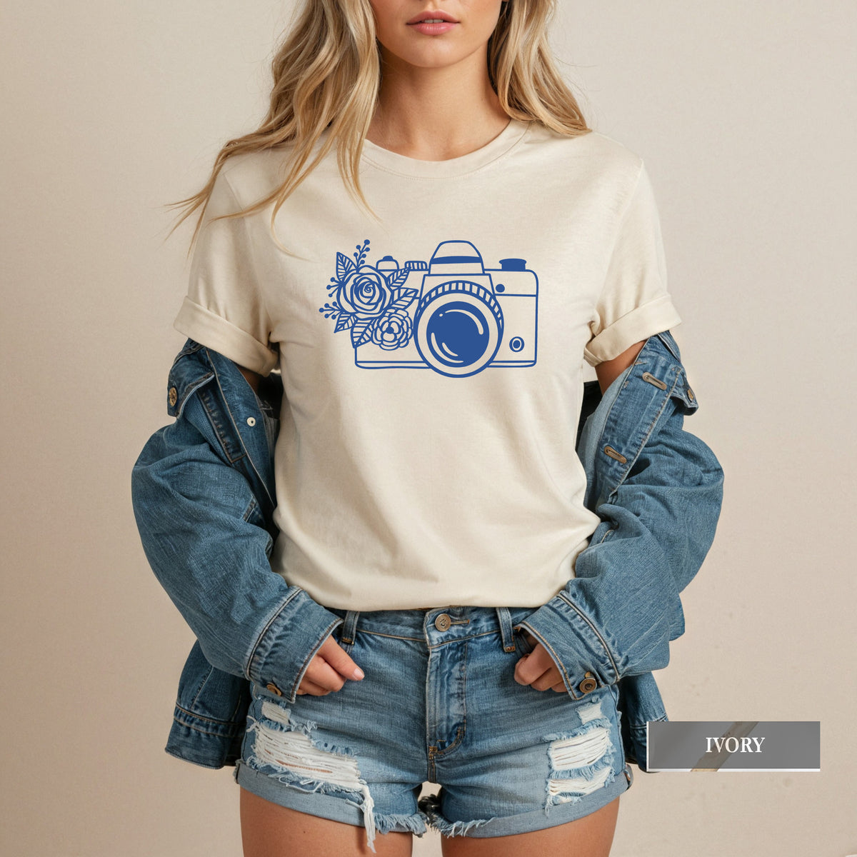 A woman wearing an ivory short-sleeve t-shirt with a blue camera and floral graphic, styled with a blue denim jacket and distressed denim shorts against a neutral beige background.