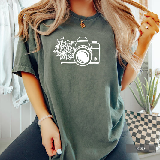 A woman wearing an oversized sage green t-shirt with a white graphic of a camera and flowers on the front, paired with black jeans in a bright indoor setting.