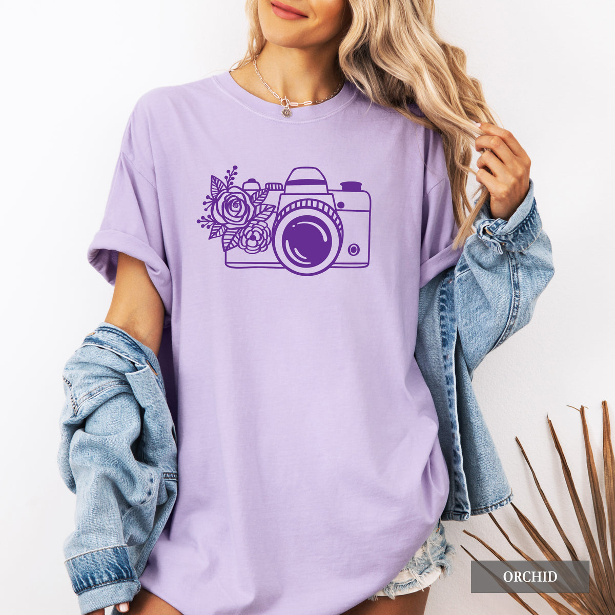 A woman wearing an oversized lavender-purple t-shirt with a purple graphic of a camera decorated with flowers. She has a light-wash blue denim jacket draped off her shoulders, and a small 'ORCHID' brand label is visible in the bottom right corner.